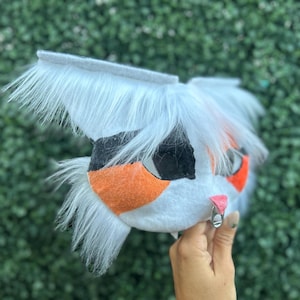 May include: A white and gray furry mask with orange and black accents. The mask has long, fluffy white fur around the ears and face. The nose is orange, and the eyes are black. A small, pink nose and a silver ring are also visible.
