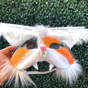 May include: A cat-themed mask with white, orange, and black fur details. The mask has large, fluffy ears and a white chin with two small teeth. A gold-colored ring is attached to the nose. The mask is held up by a white elastic strap.