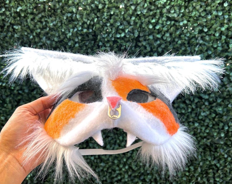 May include: A cat-themed mask with white, orange, and black fur details. The mask has large, fluffy ears and a white chin with two small teeth. A gold-colored ring is attached to the nose. The mask is held up by a white elastic strap.