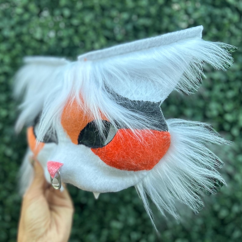 May include: A white, orange, and black cat-like mask with long white fur. The mask has a pink nose, a silver ring, and a mesh eye covering. The ears are pointed, and the fur is long and fluffy.