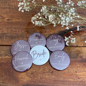 May include: Six round purple and white buttons with white text. The buttons say "Bride", "Maid of Honor", "Bridesmaid", "Bridesmaid", "Bridesmaid", and "Jessi".