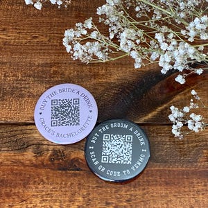 May include: Two round buttons with QR codes and text. The top button is purple with the text "Buy the Bride a Drink Grace's Bachelorette". The bottom button is black with the text "Buy the Groom a Drink Scan QR Code to Venmo".