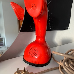 May include: A vintage, bright red telephone with a unique, curved design. The phone features a speaker and a receiver, with a coiled beige cord and a beige plug. The phone is a retro design, likely from the mid-20th century.