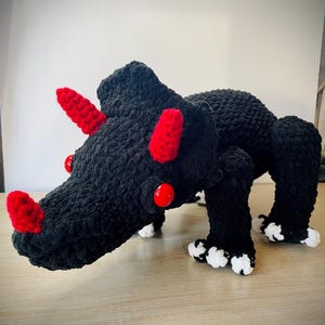 May include: A handmade crochet dinosaur in black with red horns and red eyes. The dinosaur's feet have white accents. The plush toy is sitting on a wooden surface.
