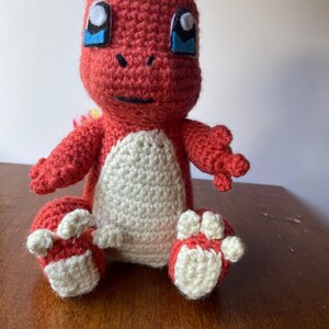 May include: A crocheted plush toy of a red and white Pokemon character with blue eyes. The toy is sitting on a wooden surface.