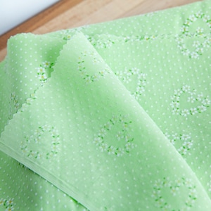 May include: Light green fabric with a repeating pattern of white flowers and small white dots. The fabric is folded over, showing the texture and design. The fabric is likely used for sewing or crafting projects.