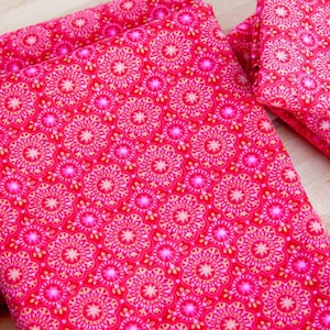 May include: Several folded pieces of fabric with a repeating floral pattern in shades of pink and red. The fabric is arranged on a light-colored wooden surface, showcasing the detailed design and color variations.