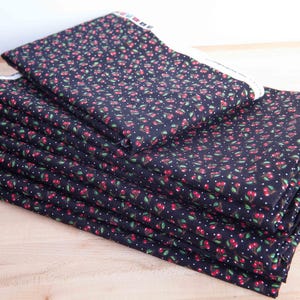 May include: A stack of dark blue fabric with a repeating pattern of red cherries, green leaves, and white dots. The fabric is neatly folded and stacked on a light brown wooden surface. The edges of the fabric have a white border.