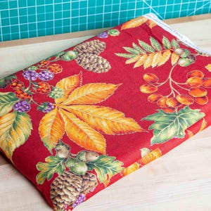 May include: A folded piece of red fabric featuring a repeating pattern of autumn-themed botanical elements, including brown pinecones, orange and yellow fall leaves, clusters of red berries, and small purple berries, with a white selvage edge.