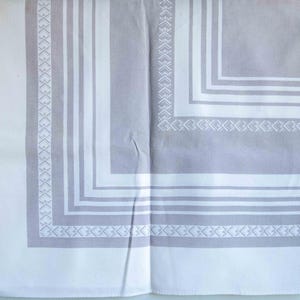 May include: A folded, rectangular tablecloth with a white and gray geometric pattern. The design features a border of repeating shapes and lines, creating a symmetrical and elegant look. The fabric appears to be cotton or linen.