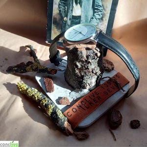 May include: A silver-toned wristwatch with a black leather band rests on a wooden stand. The watch has a white face with black hands and markers. A brown leather tag with the text "CONCLIVE" is also present. The watch is displayed on a concrete base.