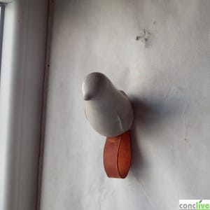 Puede incluir: Un colgador de pared con forma de pájaro de cerámica blanca con una correa de cuero marrón. El pájaro tiene un cuerpo liso y redondeado y un pico pequeño y puntiagudo. La correa de cuero está unida a la parte inferior del pájaro.