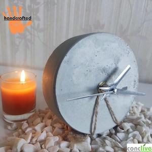 Puede incluir: Un reloj de hormigón hecho a mano con manecillas plateadas y un detalle de cuerda de yute. Una vela naranja encendida en un vaso está junto al reloj, que descansa sobre guijarros blancos. La esfera del reloj es de color gris claro.