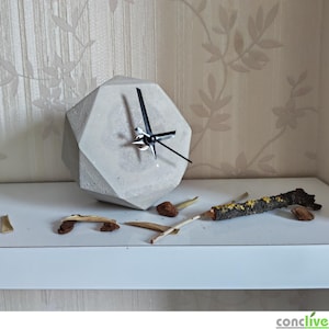 Puede incluir: Un reloj geométrico de hormigón gris con manecillas negras. Está sobre una estantería blanca, con ramitas secas y trozos de madera cerca. El fondo es un papel pintado floral. El diseño del reloj es facetado.