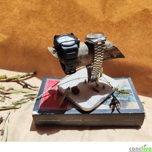 Puede incluir: Dos relojes de pulsera expuestos en un soporte hecho a mano. Un reloj es negro con una pantalla digital, el otro tiene una correa plateada y una esfera redonda. El soporte es de madera y hormigón, apoyado sobre un libro.