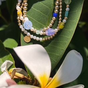 May include: Three gold beaded bracelets with colorful beads and pearls are displayed on a green leaf. The bracelets feature gold beads, small pearls, and various colorful beads in shades of blue, yellow, and purple. A small gold coin charm is also visible.