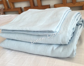 Puede incluir: Una pila de ropa de cama de lino azul claro, doblada cuidadosamente sobre una colcha blanca. El texto "Linen Legend" es visible en la ropa de cama.