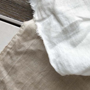 Puede incluir: Dos piezas de tela, una blanca y otra beige, se superponen. La tela blanca está en la parte superior y tiene una superficie ligeramente texturizada. La tela beige está debajo y tiene una superficie más lisa.