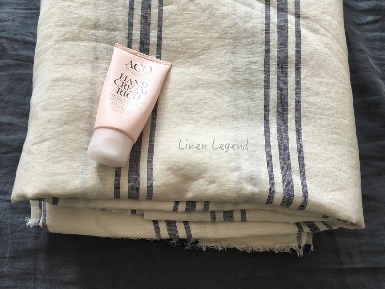 May include: A white linen fabric with blue stripes, folded in a stack. A tube of hand cream labeled "ACO Hand Cream Rich" is on top of the fabric.