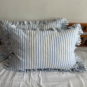 Pode incluir: Duas almofadas listradas em azul e branco com bordas com babados sobre um lençol branco. As almofadas têm uma aparência texturizada e um estilo clássico. As listras azuis são verticais e uniformemente espaçadas.