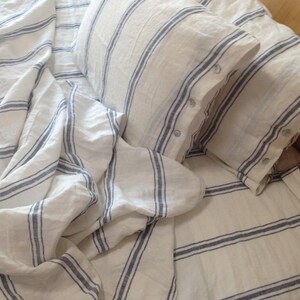 May include: White linen bedding with blue stripes. The bedding is arranged on a bed with two pillows. The pillows are also white linen with blue stripes.