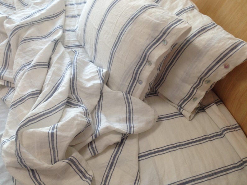 May include: White linen bedding with blue stripes. The bedding is arranged on a bed with two pillows. The pillows are also white linen with blue stripes.