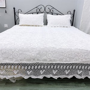 May include: White crochet bedspread with a delicate floral pattern and a scalloped edge. The bedspread is on a black metal bed frame with white pillows.