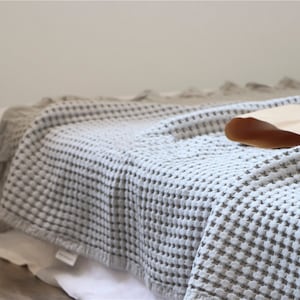 Peut inclure: Une couverture en tissu gaufré doux de couleur gris clair. La couverture est pliée et drapée sur un lit, une partie de la couverture dépassant du bord.