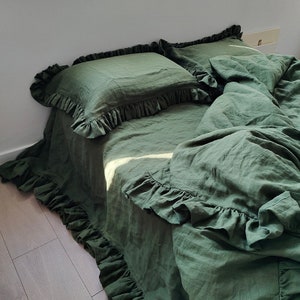 May include: Dark green linen bedding set featuring ruffled detailing. The set includes a duvet cover and pillowcases. The bedding is displayed on a bed, with sunlight highlighting the fabric. The overall style is rustic and comfortable.