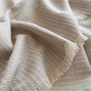 May include: A close-up of a beige waffle weave throw blanket with a white fringe. The blanket is soft and textured, and the fringe adds a touch of elegance.