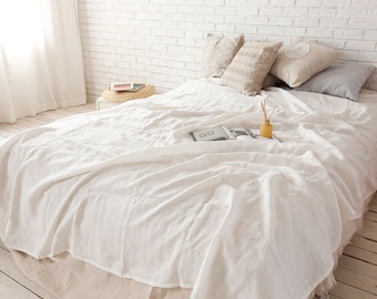 Könnte beinhalten: Weiße Bettwäsche aus Leinen auf einem Bett mit einer weißen Backsteinwand dahinter. Das Bett ist mit einer weißen Leinenbettdecke und mehreren Kissen in verschiedenen Beige- und Grautönen bezogen. Ein kleiner Holz-Beistelltisch mit einem Buch und einem Dekorationsgegenstand ist im Hintergrund zu sehen.