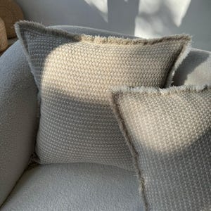 May include: Two beige throw pillows with a textured woven fabric and frayed edges. The pillows are on a light beige couch.