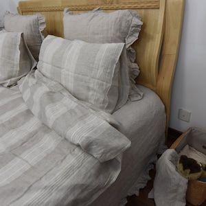 May include: A bed with a white and gray striped linen duvet cover and matching pillowcases. The bed has a wooden headboard with a carved design. There is a wicker basket on the floor next to the bed.