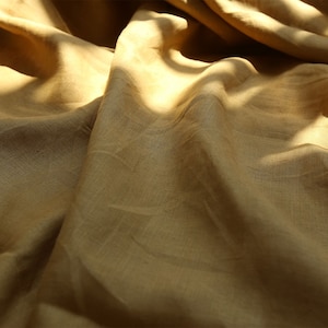 Peut inclure: Gros plan sur un tissu drapé de couleur brun doré chaud. Le textile a une texture subtile et est éclairé par une lumière douce, créant des ombres et soulignant les plis. L'image met en valeur le drapé et le flux naturels du matériau.