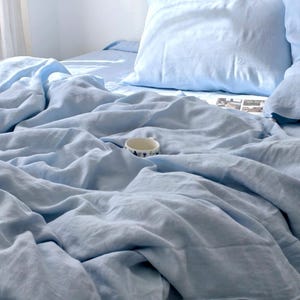 May include: A light blue bedding set featuring a rumpled duvet cover and a matching pillowcase. A small, white ceramic mug with a dark blue patterned rim sits nestled within the folds of the soft, textured fabric on the bed.