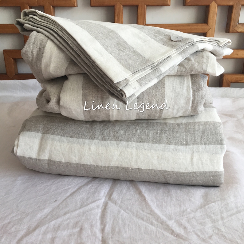 May include: A stack of three grey and white striped linen bedding sets. The top set has a button closure. The bedding is folded and arranged on a white surface. The text "Linen Legend" is visible on the bedding.