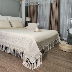 May include: Cream-colored bedspread with a geometric lace pattern and fringe trim. The bed is made with matching pillowcases and a decorative pillow. A cup and book are on the bed.