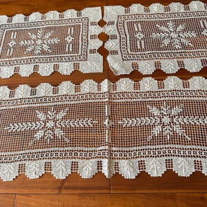 Puede incluir: Camino de mesa de encaje crema con diseño de copo de nieve. El camino de mesa presenta un borde festoneado con recortes en forma de hoja y patrones florales intrincados. Colocado sobre una superficie de madera.