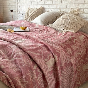 Op de afbeelding: Een roze en wit gepatroneerde deken met een wit gehaakt kussen en een beige kussen op een bed met een witte bakstenen muur op de achtergrond.