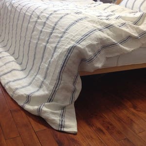May include: A white and blue striped linen bed sheet with a blue stripe pattern. The sheet is draped over a bed frame and the edges are hanging down.