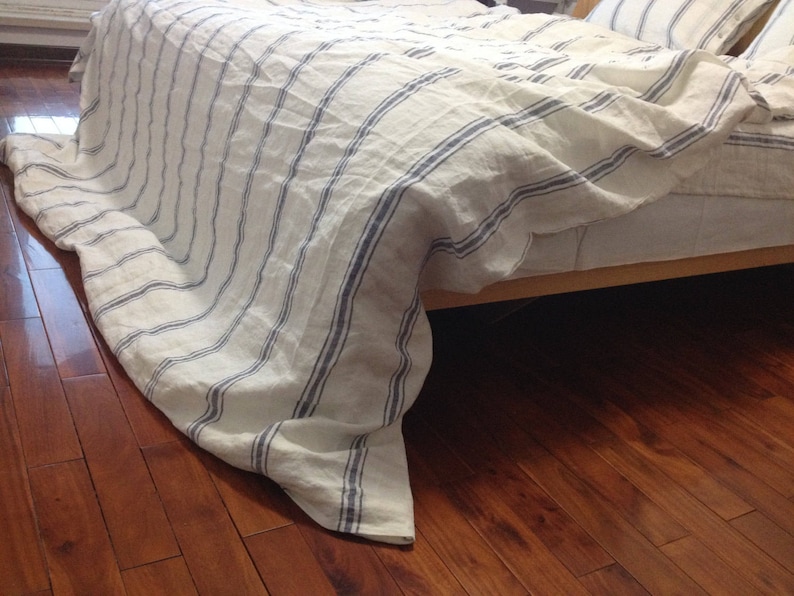 May include: A white and blue striped linen bed sheet with a blue stripe pattern. The sheet is draped over a bed frame and the edges are hanging down.