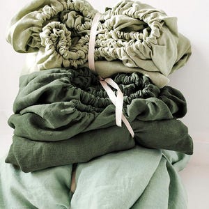 May include: A stack of three fitted bed sheets in shades of green. The top sheet is a light green, the middle sheet is a darker green, and the bottom sheet is a light green. The sheets are tied together with a white ribbon.