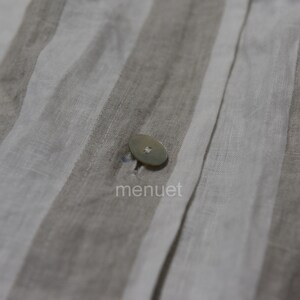 May include: Close-up of a white button sewn onto a beige and white striped fabric. The fabric has a linen texture.