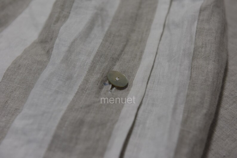May include: Close-up of a white button sewn onto a beige and white striped fabric. The fabric has a linen texture.