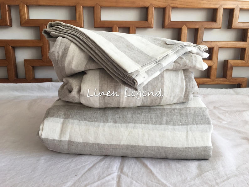 May include: A stack of three grey and white striped linen bedding sets. The bedding is folded and arranged on a white bed. The text "Linen Legend" is visible on the middle set.