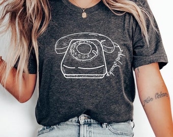 Può includere: Una t-shirt color grigio scuro melange con un disegno bianco di un telefono a disco e il testo "componi i tuoi sogni".