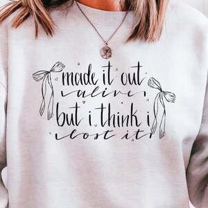 May include: Ash-colored sweatshirt with the text "made it out alive, but I think I lost it." The design includes two bow illustrations. The word "Ash" is printed on a banner at the bottom of the image. The sweatshirt is a casual style.