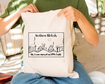 Puede incluir: Una bolsa de lona blanca con una ilustración en blanco y negro de un pueblo nevado con el texto "Northern Altitude" y "Oh, I was raised on a little light".