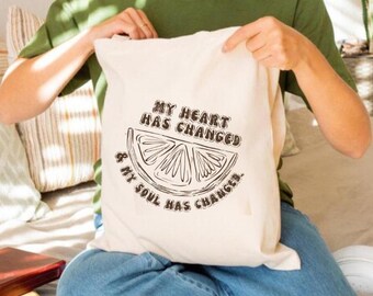 Puede incluir: Una bolsa tote de color crema con el texto "MY HEART HAS CHANGED & MY SOUL HAS CHANGED" y un gráfico de una rodaja de limón. La bolsa la sostiene una persona que lleva una camiseta verde y vaqueros azules. El fondo incluye plantas y libros.