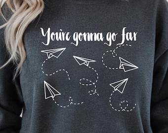 Puede incluir: Sudadera gris jaspeado oscuro con el texto "You're gonna go far" y gráficos de aviones de papel blancos. La sudadera de cuello redondo tiene mangas largas. Las palabras "Dark Heather" están impresas en la parte inferior.
